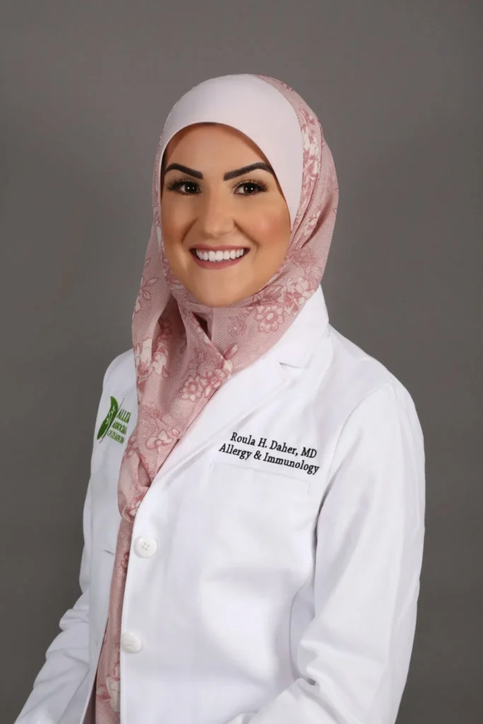 Female doctor in a white coat and pink hijab, smiling against a gray background.