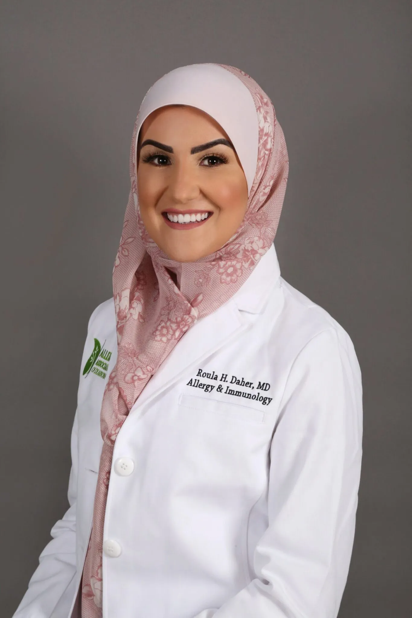 Female doctor in a white coat and pink hijab, smiling against a gray background.